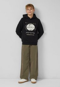 Ragazzo in piedi contro un muro, che indossa un hoodie nero con testo grafico e pantaloni larghi olivastro, abbinati a sneakers bianche.