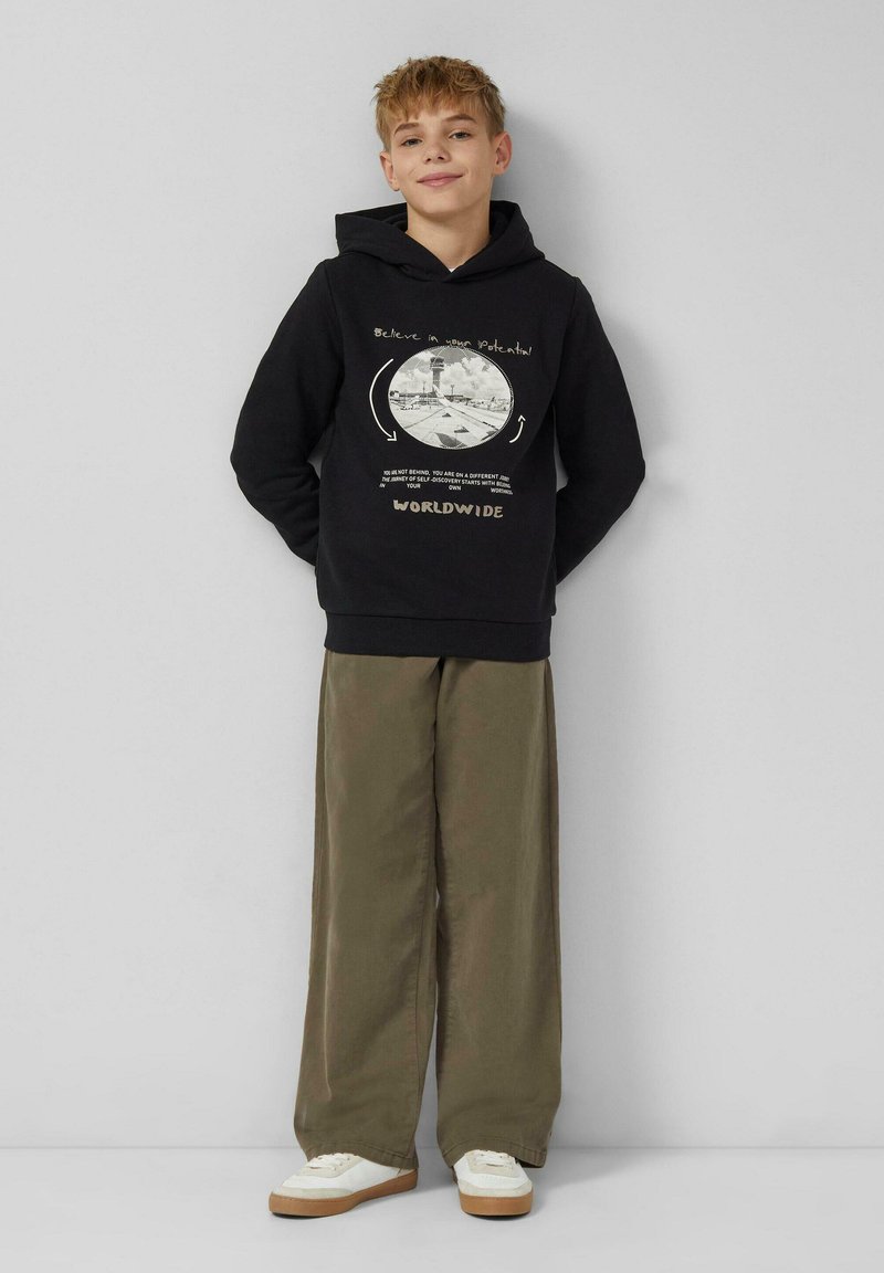 Ragazzo in piedi contro un muro, che indossa un hoodie nero con testo grafico e pantaloni larghi olivastro, abbinati a sneakers bianche.