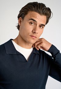 Granatowy dzianinowy sweter z podwiniętym kołnierzykiem i białą koszulą pod spodem. Tekstura materiału wydaje się być miękka i lekka, z dopasowanym krojem.