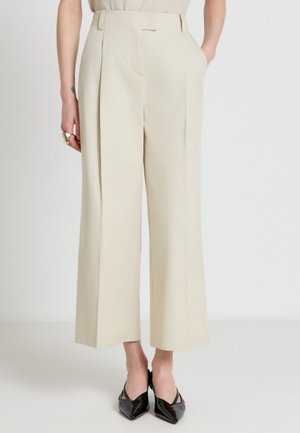 Pantaloni beige a gamba larga, tagliati sopra la caviglia, indossati con décolleté nere a punta, mani parzialmente nelle tasche, su uno sfondo chiaro e uniforme.