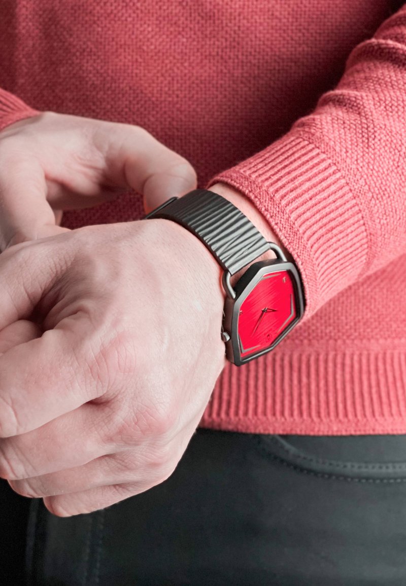 Hexagonal ur med en lys rød urskive, sort rillet silikonearmbånd og sølv hardware. Båret på et håndled med en tekstureret rød sweater.