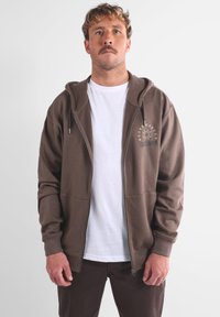 Felpa con zip marrone, con tasca frontale, cappuccio con cordoncino e una grafica bianca sul lato sinistro del petto. Indossata sopra una maglietta bianca e pantaloni marroni.