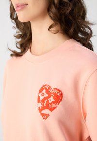 Lys rosa hettegenser med rund hals, med et rødt hjertegrafikk og abstrakte former, laget av mykt bomullsmateriale.
