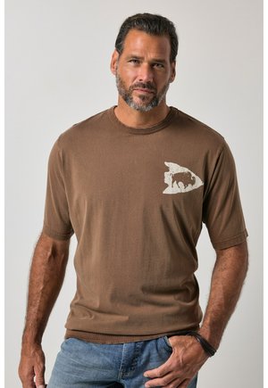 Mann mit kurzen dunklen Haaren und Bart, trägt braunes T-Shirt mit Bisongrafik und blaue Jeans, posiert vor neutralem Hintergrund.