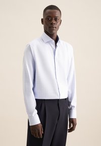 Camicia azzurra chiara con un sottile motivo a griglia, maniche lunghe e colletto classico. Abbinata a pantaloni scuri su misura.