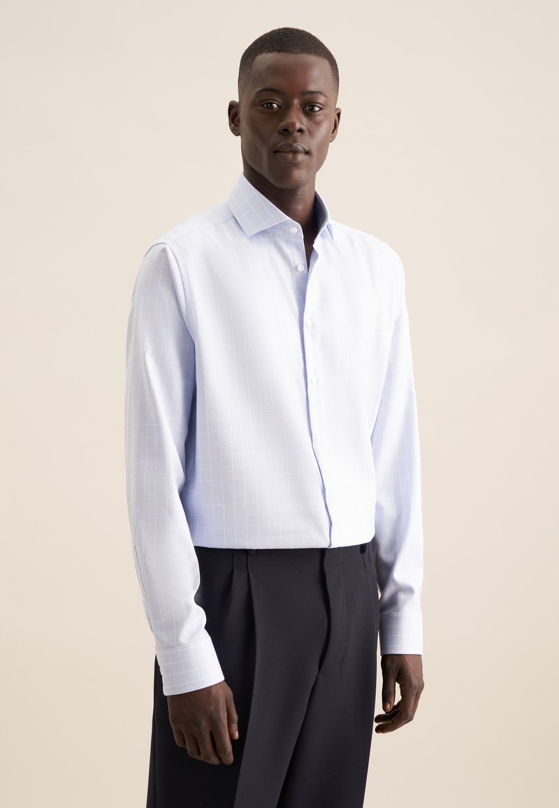 Camicia azzurra chiara con un sottile motivo a griglia, maniche lunghe e colletto classico. Abbinata a pantaloni scuri su misura.