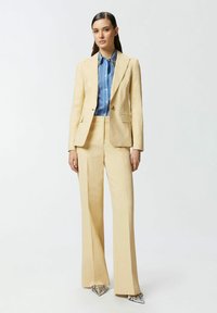 Svijetložuti odijelo po mjeri s prilagođenim sakoom i širokim hlačama, uparen s plavom prugastom košuljom i srebrnim cipelama sa šiljatim vrhom.