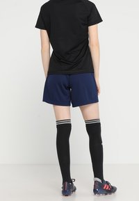 Camisa deportiva negra de manga corta, pantalones cortos azules, medias negras hasta la rodilla con rayas blancas y botas de fútbol negras con acentos rojos.