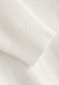 Primo piano del tessuto di un maglione bianco, con dettaglio del polsino a costine e texture morbida, che mostra una maglia liscia e uniforme e una palette di colori neutri.