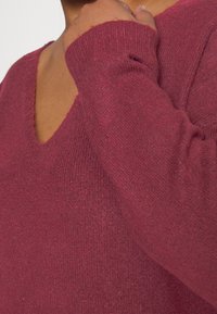 Burgunderfarbener Strickpullover mit V-Ausschnitt und langen Ärmeln. Der Stoff hat eine weiche Textur mit leichtem Stretch und sichtbarem Strickmuster.