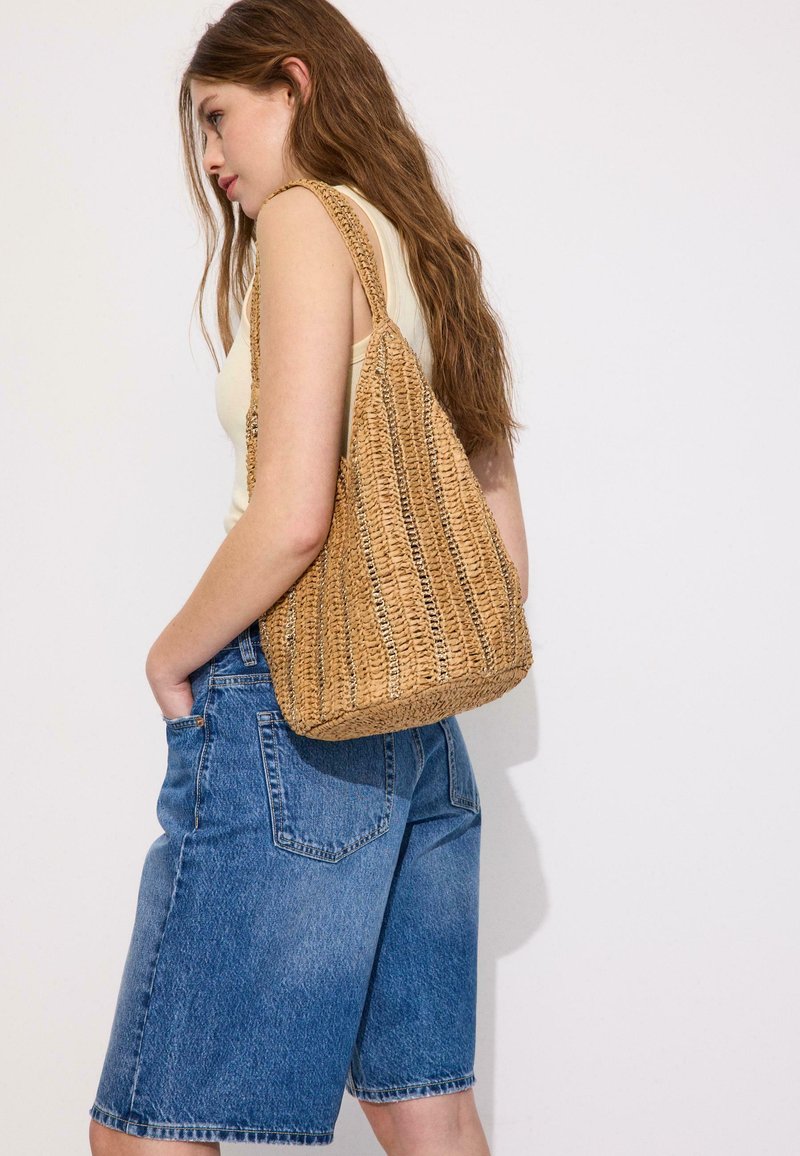 Jeune femme aux longs cheveux bruns portant un top sans manches crème, un short en jean bleu et un grand sac d'épaule tressé en paille.