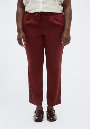 Osoba o ciemnej karnacji ubrana w bordowe spodnie ze ściągaczem i pasujące mokasyny, stojąca na tle jednolitego białego tła.