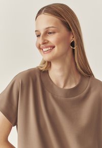 Braunes kurzärmliges Oberteil mit rundem Ausschnitt und glatter Textur. Goldene und schwarze asymmetrische Ohrringe mit flüssigem Design.