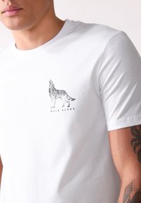 Camiseta de algodón blanca con un gráfico negro de un lobo aullando y el texto "WALK ALONE" debajo. Mangas cortas y cuello redondo.
