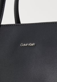 Black leather tote bag featuring a silver "Calvin Klein" logo, smooth texture, and meticulous stitching along the edges.
