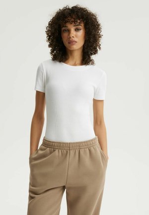 Kobieta o kręconych włosach, ubrana w dopasowany biały krótki top z krótkim rękawem i luźne beżowe spodnie, z rękami w kieszeniach, stojąca na jednolitym tle.