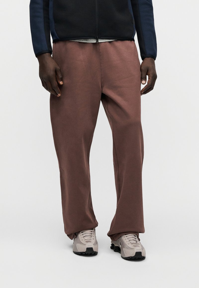 Braune, locker sitzende Jogginghosen mit glatter Textur, kombiniert mit hellgrauen Sportschuhen mit schwarzen Akzenten und Schnürsenkeln.