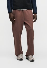 Rudos laisvai priglundančios sportinės kelnės su lygiu paviršiumi, derinamos su šviesiai pilkomis sportinėmis batelėmis, kuriose yra juodos detalės ir raišteliai.