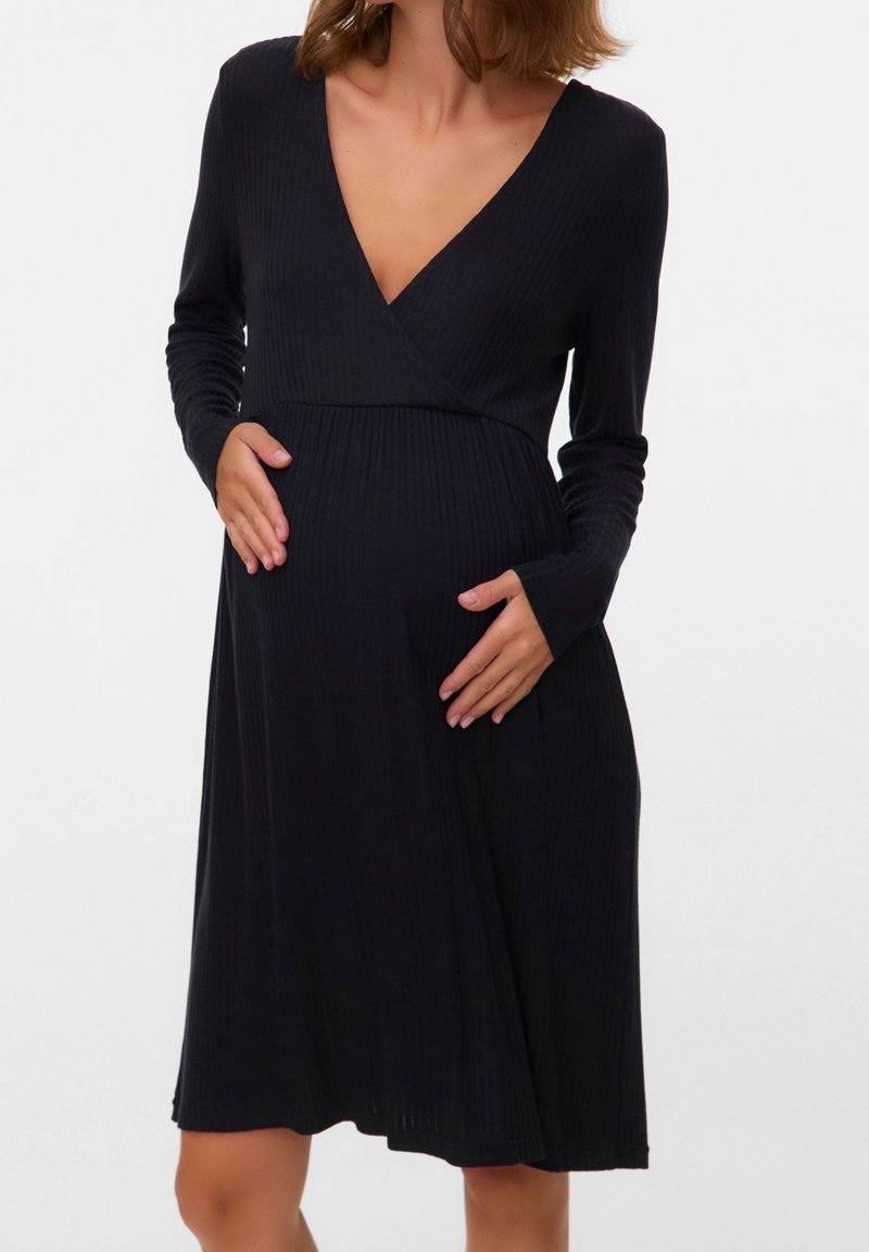 Femme enceinte portant une robe noire côtelée manches longues avec un décolleté en V, tenant délicatement son ventre avec ses deux mains.