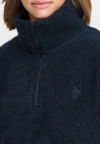 Marineblå fleece-pullover med høy krage og halv-zip-lukking, med teksturert stoff og en diskret merkevarelogo på brystet.