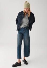 Navy quilted bomber jacket, grey sweater, light green ribbed beanie, wide-leg blue jeans, and black lace-up shoes. Plain background.