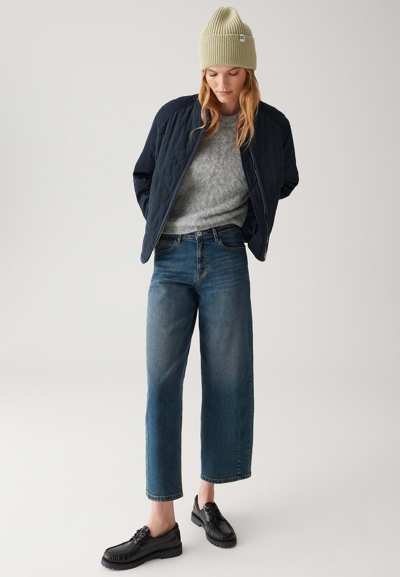 Navy quilted bomber jacket, grey sweater, light green ribbed beanie, wide-leg blue jeans, and black lace-up shoes. Plain background.