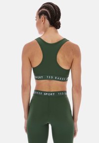 Žalia sportinė liemenėlė su apvalia nugara ir prekės ženklo juosta. Derinama su atitinkančiomis aukšto liemens antblauzdėmis su tuo pačiu logotipu. Lygus tekstūra.