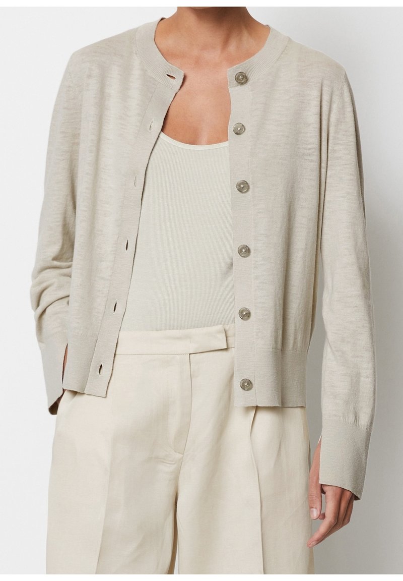 Beżowy kardigan z okrągłym dekoltem, zapinany na guziki z przodu, z długimi rękawami. Miękka tekstura i subtelny połysk w jasnych, neutralnych tonach.