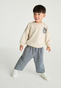 Lichtbeige katoenen sweatshirt met een kleine blauwe grafiek; marineblauwe en witte gestreepte loszittende broek; witte sneakers; kind dat staat en glimlacht.