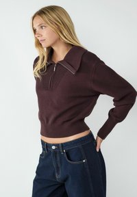 Dunkelbrauner Strickpullover mit hohem, geripptem Kragen und halbem Reißverschluss. Cropped-Design mit geripptem Saum, kombiniert mit dunklen Denim-Jeans.