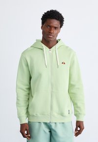Zelena zip-up dukserica s prednjim džepom, kapuljačom s ušicom za zatezanje i izvezenim logotipom na prsima. Glatka tekstura i rebrasti manžeti.