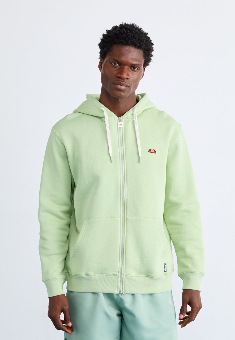 Zelena zip-up dukserica s prednjim džepom, kapuljačom s ušicom za zatezanje i izvezenim logotipom na prsima. Glatka tekstura i rebrasti manžeti.