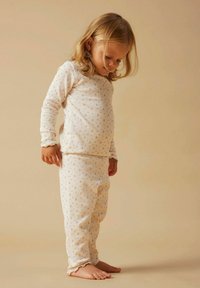 Młode dziecko o blond włosach, ubrane w kremową, długą koszulę i spodnie w małe kwiatowe wzory, stoi boso na beżowej podłodze.