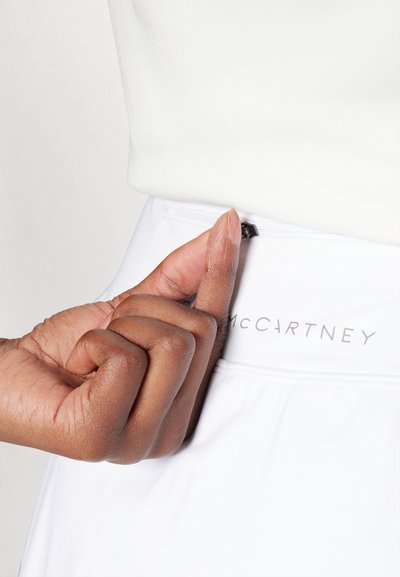 Fehér atlétikai rövidnadrág sima felülettel, kis fekete cipzárral és ezüst "McCartney" logóval a derékpánton. Egy kéz fogja a cipzárt.