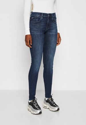 Jeans de mezclilla azul oscuro de corte ajustado con cinco bolsillos, talle medio y un leve desgastado. Combinados con zapatillas negras y plateadas.