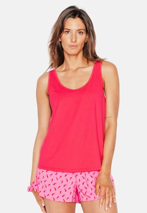 Femme aux cheveux bruns portant un débardeur rouge sans manches et un short rose à motif de piments rouges, debout devant un fond blanc.