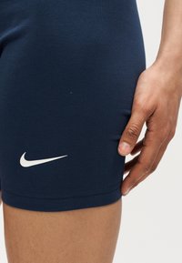 Κοντινό πλάνο ενός χεριού που ακουμπά κοντά στο τελείωμα ενός σκούρου μπλε αθλητικού σορτς με λευκό λογότυπο Nike.