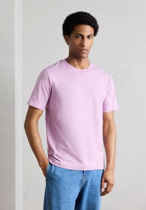 Νεαρός με κοντά σγουρά μαλλιά, φορώντας μωβ ανοιχτό κοντομάνικο μπλουζάκι και μπλε τζιν, στέκεται μπροστά σε απλό ανοιχτό γκρι φόντο.