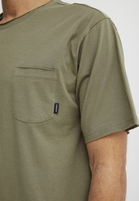 Olive green cotton T-shirt with short sleeves, a chest pocket, and a visible brand label on the side. Smooth texture and relaxed fit.