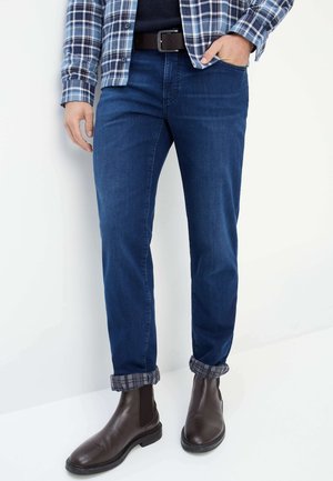 Dark blue denim jeans with a straight cut, featuring rolled cuffs revealing a plaid lining. Paired with brown leather ankle boots.