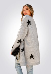 Cárdigan gris de gran tamaño con patrones de estrellas negras, textura de punto y bolsillos laterales. Combinado con un bolso negro.
