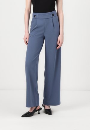 Pantalons large bleu clair à taille haute, avec une fermeture à double bouton et des plis avant, en tissu lisse. Escarpins noirs à bride arrière.