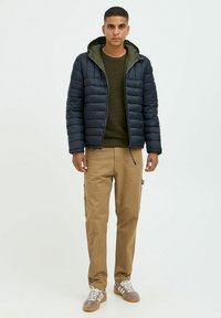 Mørkeblå pufferjakke med grøn hætte, olivengrøn striktrøje, khaki cargo-bukser og gråbrune sneakers med hvide snørebånd. Afslappet udendørsbeklædning.