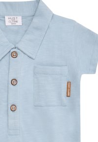 Polo shirt azzurro chiaro realizzato in tessuto textures, con colletto, tre bottoni in legno, taschino sul petto e una piccola etichetta marrone sul lato.