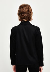 Blusa nera a maniche lunghe con colletto abbottonato, texture morbida e orlo leggermente curvato. Presenta spalle strutturate e polsini aderenti.