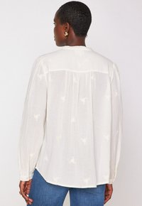 Persona con capelli corti che indossa orecchini d'oro, una blusa bianca a maniche lunghe e morbida con motivi floreali sottili e jeans blu, mostrata di spalle.