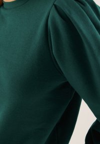 Ciemnozielony, teksturowany sweter z okrągłym dekoltem i bufkami. Zbliżenie ukazujące detal rękawów i dopasowane body.