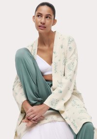 Cardigan a maglia color crema con motivi a conchiglia e sole verdi. Abbinato a pantaloni larghi verde menta e un crop top bianco. Stile casual, vestibilità rilassata.