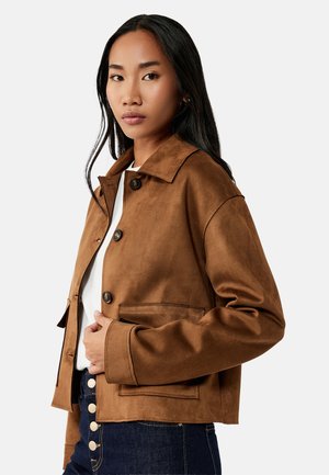 Mujer con cabello largo y negro que lleva una chaqueta marrón abotonada sobre una camiseta blanca y vaqueros azul oscuro con botones visibles.