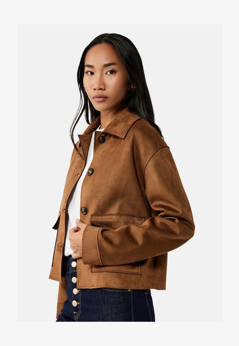 Frau mit langen schwarzen Haaren, die eine braune Jacke mit Knöpfen über einem weißen Top und dunkelblauen Jeans mit sichtbaren Knöpfen trägt.